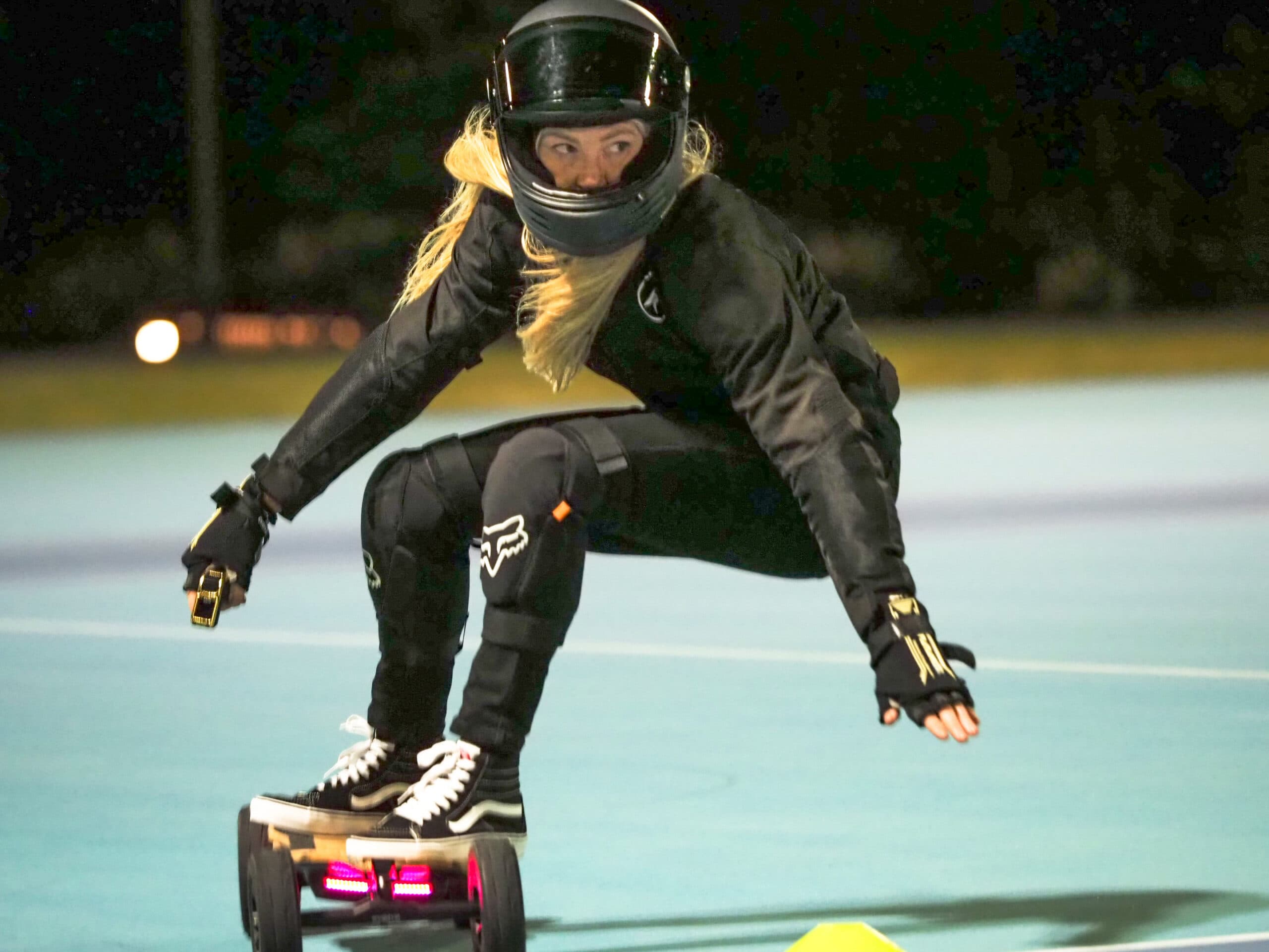 Bianca Criaco crouched low in racing gear on an electric skateboard mid-race at night.